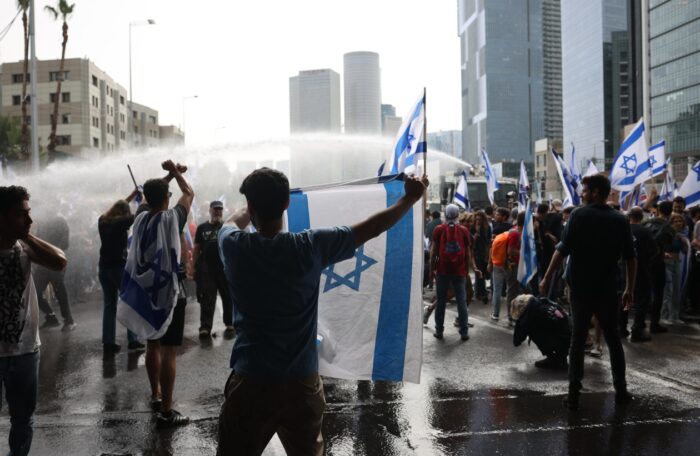 Man sieht von hinten eine Gruppe von Menschen, die mit Wasser beschossen werden. Einer hält eine Israel-Flagge hoch.