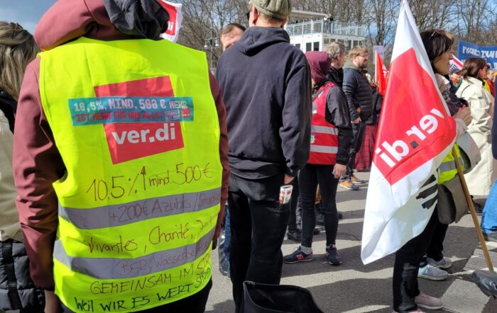 Man sieht mehrere Personen, einer hat eine gelbe verdi Weste an, eine andere trägt eine verdi Fahne.