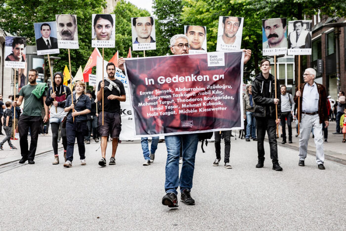Das Bild zeigt die Spitze der Demonstration zum Gedenken an die zehn Opfer des NSU, im Juli 2018 in Hamburg. Im Vordergrund läuft ein Mann, der ein Banner hält, auf dem die Namen der NSU-Opfer zu lesen sind. Direkt hinter ihm bilden zehn Menschen eine Reihe. Jede Person hält ein Schild in der Hand auf dem je ein NSU-Opfer zu sehen ist.