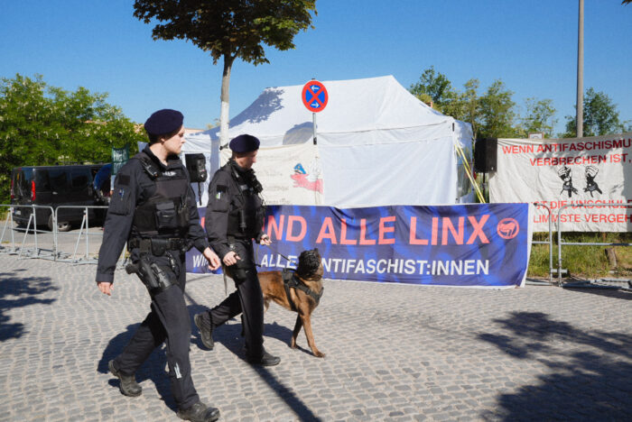 Vor einem Solidaritätsplakat für Antifaschist*innen laufen Polizisten mit einem Schäferhund Patrouille.