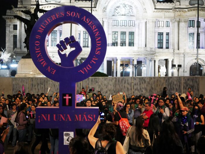 Eine Gruppe Menschen protestiert. In der Mitte steht ein Kreis mit einem Kreuz unten in violett. In der Mitte des Kreises ist eine Faust - ein kämpferisches Zeichen gegen Gewalt an Frauen.