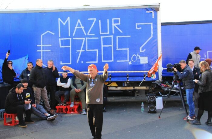 Ein Mann tanzt auf einem Parkplatz vor einem LKW, im Hintergrund weitere Männer, die teils auf Getränkekisten sitzen, und ein Filmteam. Auf der blauen LKW-Plane steht in großen weißen Buchstaben "Mazur € 97.585?"