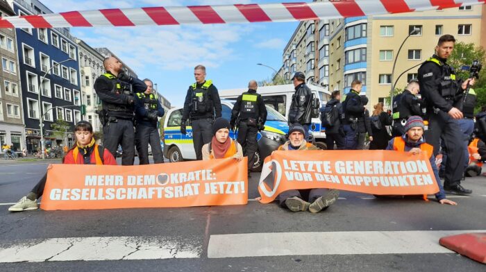 Vier Menscheni n Warnwesten sitzen auf der Straße und halten Transparente mit den Aufschriften "Mehr Demokratie: Gesellschaftsrat jetzt" und "Letzte Generation vor den Kipppunkten". Dahinter stehen Polizist*innen, im Vordergrund ein Absperrband.