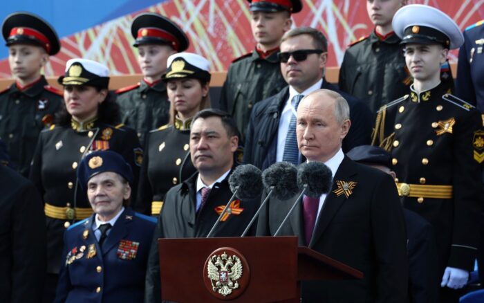 Putin steht zwischen Uniformierten an einem Rednerpult und schaut ernst drein