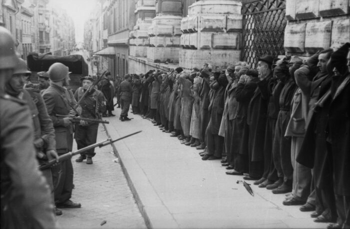 Eine historische Aufnahme: Rechts eine Reihe von Zivilistinnen und Zivilisiten, die mit erhobenen Händen an einer Mauer stehen. Davor italienische und deutsche Soldaten, die sie bewachen.
