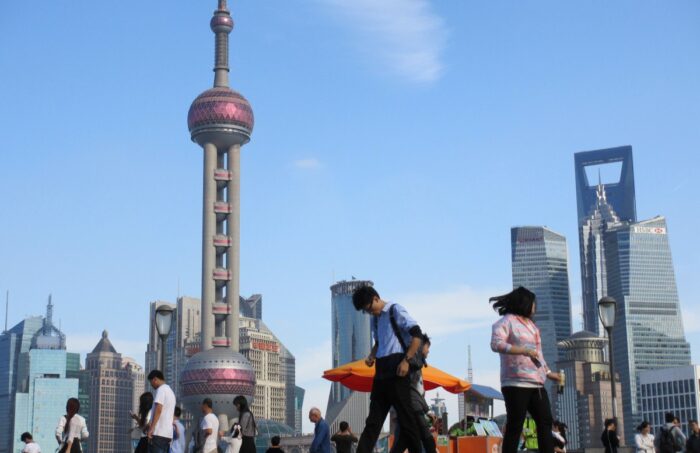 Menschen laufen auf einer Straße vor der Skyline von Shanghai entlang. Niemand sieht in die Kamera