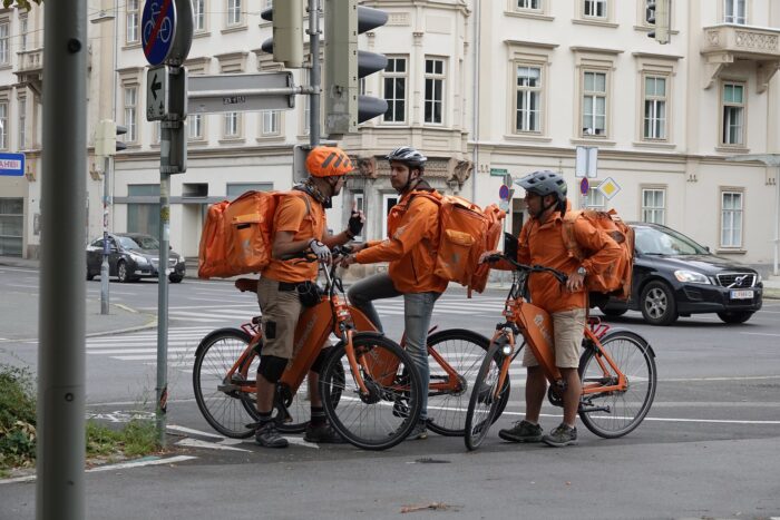 Drei Lieferando-Fahrer*innen treffen sich während ihrer Arbeit an einer Straßenecke und unterhalten sich.