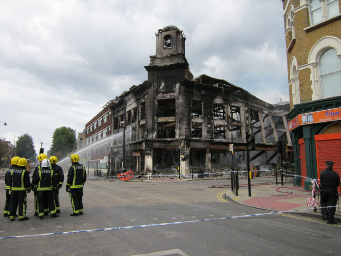 Eine Gruppe Feuerwehrleute hält den Strahl eines Wasserschlauches auf ein fast ausgebranntes Haus an einer Kreuzung, auf dem Haus ein repräsentatives Türmchen