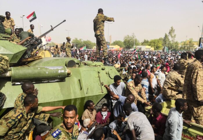 Bild einer Demonstration mit Tausenden von Zivilist*innen, einige schwenken Flaggen des Sudans. Vorne auf dem Bild ist ein Militärpanzer zu sehen, Soldaten beobachten die Szene.