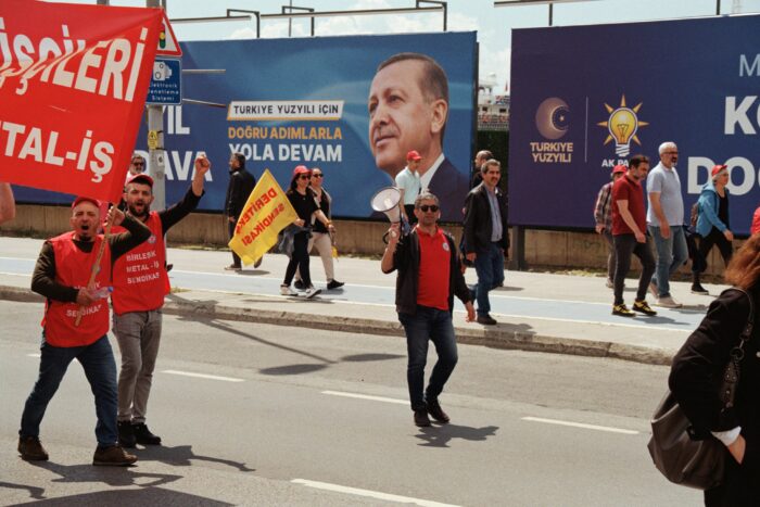 Eine Person mit rotem T-Shirt, in der rechten Hand ein Megaphon, neben ihr andere Personen in roten Westen, die ein Banner tragen, im Hintergrund ein Wahlplakat mit einem Mann im Anzug