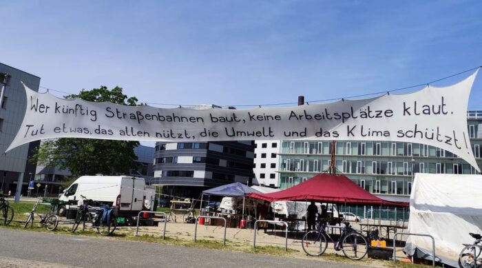 Ein großes Transparent, auf dem steht: "Wer künftig Straßenbagnen baut, keine Arbeitsplätze klaut. Tut etwas, das allen nützt, die Umwelt und das Klima schützt." Im Hintergrund Zelte eines Camps.