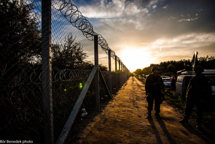 Im Abendlicht ein Zaun mit Stacheldraht, rechts daneben auf einem Weg zwei Soldaten im Hintergrund Bäume und Autos.