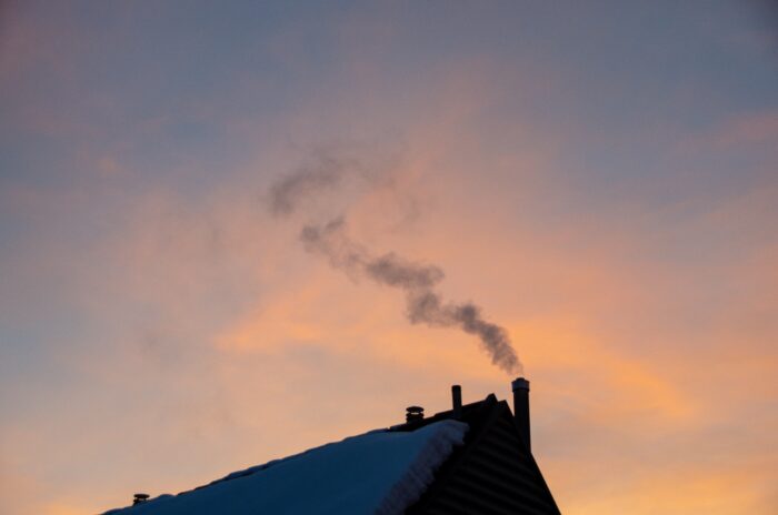Teil eines schneebedeckten Daches mit Schornstein, aus dem Rauch kommt.