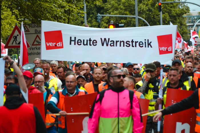 EIne Demonstration zieht durch Hamburg, die Menschen auf dem Bild tragen ein ver.di-Transparent mit der Aufschrift "Heute Wanstreik". Einige haben rote Schilder mit Buchstaben drauf, und klopfen mit Holzstangen auf die Schilder. Es ist der Hafenstreik, Sommer 2023.