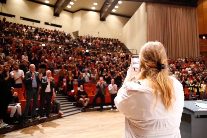 Bei der Streikkonferenz in Bochum hält Anuschka Mucha, die von hinten zu sehen ist, ihre Handykamera auf das Publikum gerichtet. Nach ihrem Bericht über einen elfwöchigen Streik an den Unikliniken in NRW gab es standing ovations.