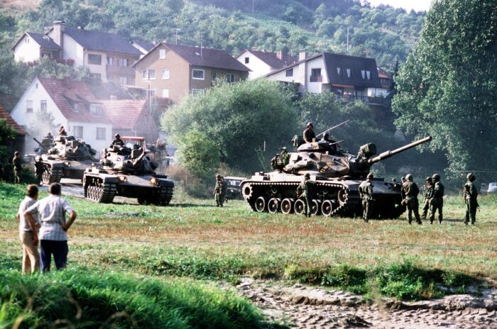 Drei Panzer überqueren ein Feld, im Vordergrund einie Menschen in ziviler Kleidung, die dabei zu schauen. Im Hintergrund eine Wohnsiedlung mit rot geklinkerten Häusern.
