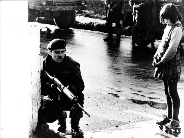 Ein Soldat kniet an der linken Bildseite an einer Häuserecke und hat ein Sturmgewehr im Anschlag. Rechts im Bild ein Mädchen auf Rollerskates, das ihn anstarrt.