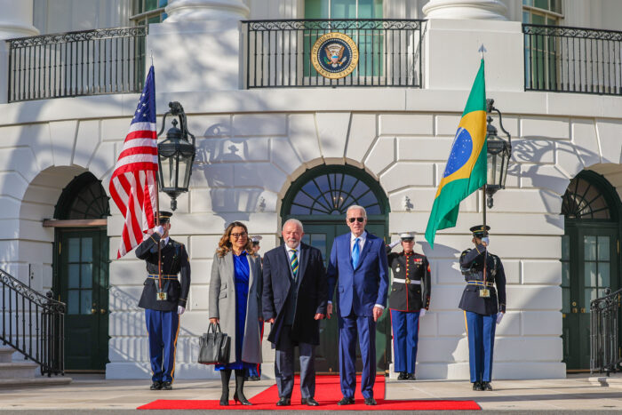 Der brasilianische Präsident Lula und der US-Präsident Biden stehen neben nebeneinander aus einem roten Teppich und gucken geradeaus in die Kamera. Neben Lula steht noch eine Frau, Rosângela Lula da Silva. Im Hintergund halten SOldaten die brasilianischen und US-Amerikanische Flagge in die Höhe.