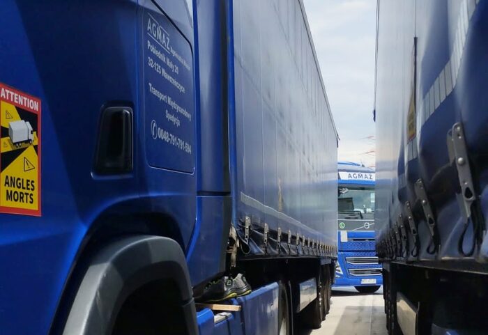 Blick durch zwei eng nebeneinander geparkte blaue LKW, durch die man das Fahrerhaus eines dritten mit der Aufschrift "Agmaz" sieht