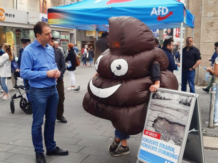 Eiin großer Haufen Scheiße unterhält sich an einem AfD Wahlkampfstand mit einem Mann.