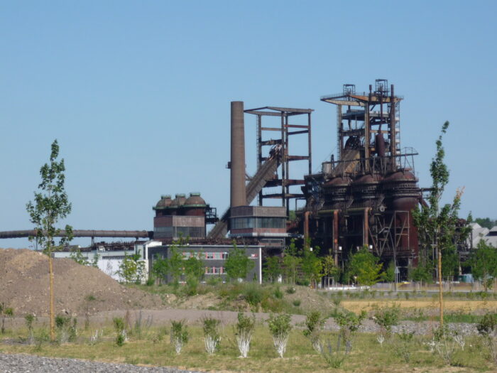 Alte, rostige Industrietürme ragen in den Himmel, im Vordergrund sind Gräser und Pflanzen zu sehen