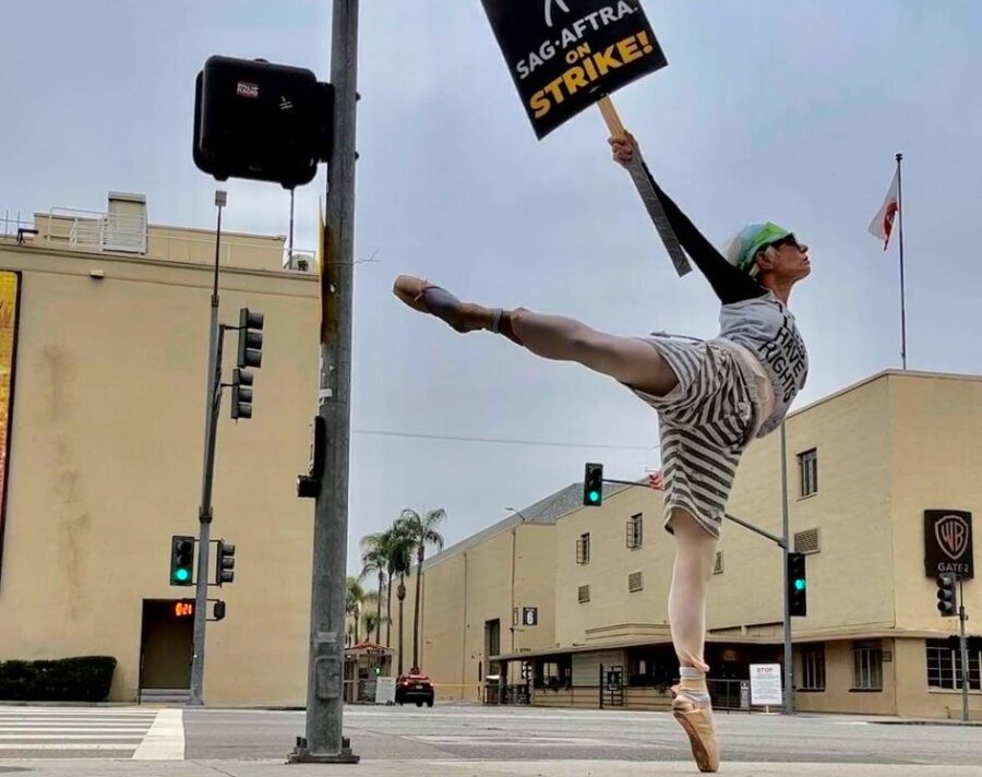 Eine Tänzerin in eleganter Poste mit abgespreiztem Bein hält ein Schld mit der Aufschrift "SAG-AFTRA STRIKE!", im Hintergrund ein Gebäude mit Warner Bros Schild