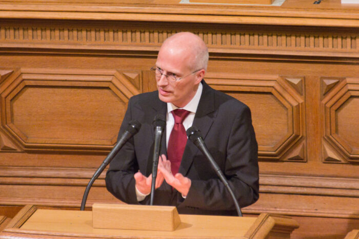 Der erste Bürgermeister der Stadt Hamburg, Peter Tschentscher, steht an einem Rednerpult vor einer holzbetäfelten Wand, mutmaßlich im Hamburger Rathaus.