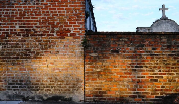 Eine Backsteinmauer, auf die die Sonne fällt, rechts hinter der Mauer ist ein Kreuz zu erkennen