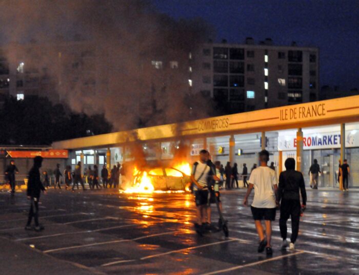 Ein brennendes Auto steht auf einem Parkplatz, dahinter ein Supermarkt mit weißem Schild, um das Auto stehen Jugendliche