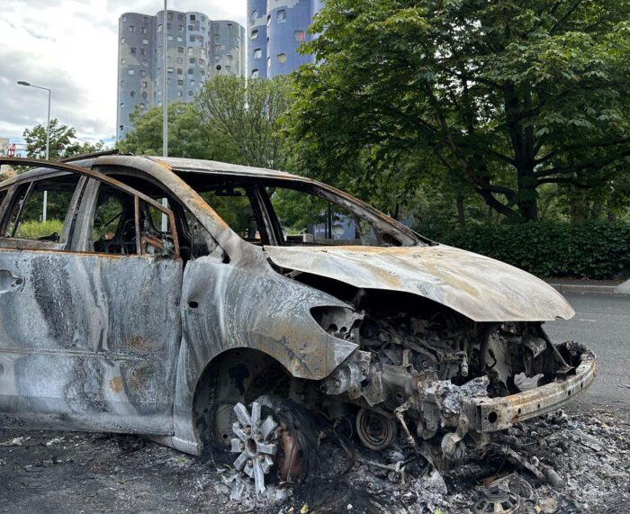 Ein ausgebranntes Auto, im Hintergrund ein Baum, dahinter ein Hochhaus