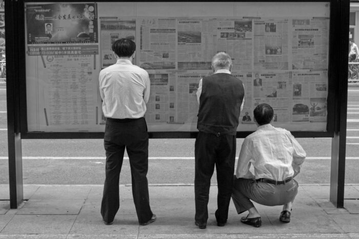 Drei Männer stehen in Shanghai vor einem Glaskasten, in dem Zeitungen aufgehängt sind. Sie lesen. Das Bild ist schwarz-weiß.