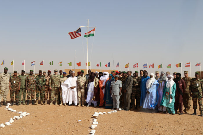 Gruppenphoto. Eine Gruppe von Menschen mit und ohne Uniform posieren für ein Bild. Im Hintergrund sind die Fahnen vieler Länder zu sehen. In der Mitte hinten ist ein großes Fahnenmast mit den Flaggen Nigers und der USA.