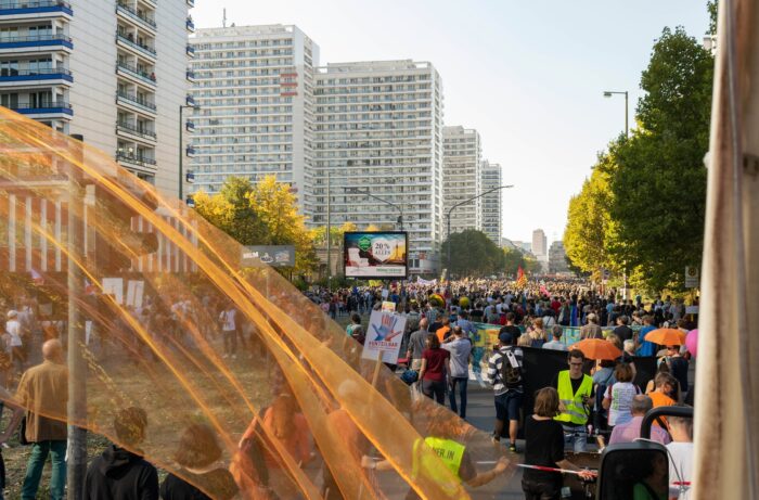 Demonstration auf einer breiten, von Hochhäusern gesäumten Straße, von links hängt ein halbdurchsichtges orangefarbenes Tuch über dem Bild