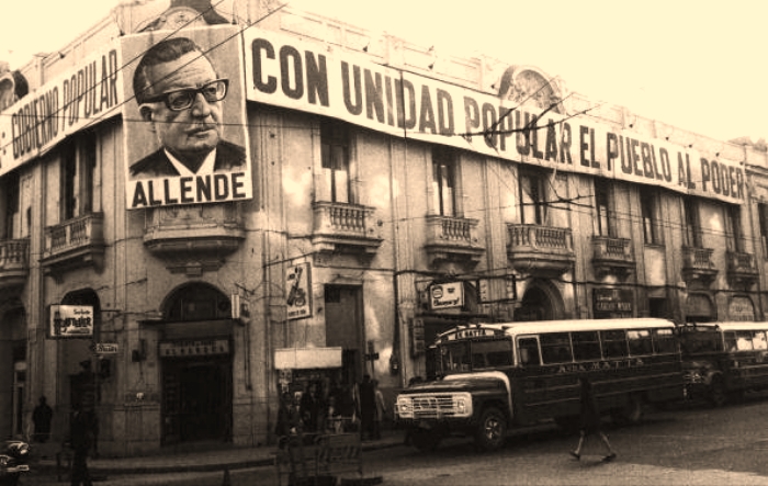 Photo einer Straßenecke. Ein altes Gebäude mit großen Bannern mit spanischer Aufschrift "für die Volkseinheit alle Macht dem Volk". An der Ecke ein Porträt von Salvador Allende.