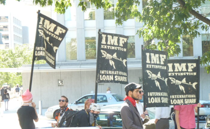 Ein paar Demonstranten halten Schilder in die Höhem die das Skelet eines Haies zeigen. Die Aufschrift sagt "IWF: Internationale Kredithaie"