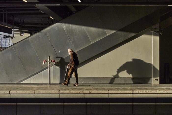 Ein alter Mann mit Maske geht gebeugt einen Bahnsteig entlang und wirft im niedrig stehenden Sonnenlicht einen langen Schatten