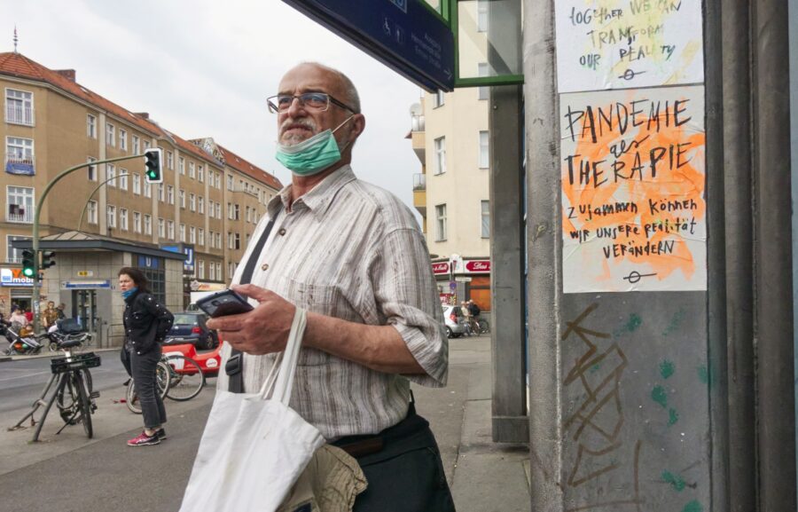 Straßenszene: Foto eines Mannes mit Maske unter dem Kinn. neben ihm klebt an der Wand ein Plakat mit der Aufschrift "Pandemie als Therapie"