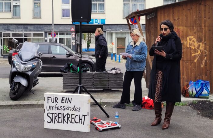 Eine Frau mit Sonnenbrille und dunklem Mantel spricht auf einer Art Parkplatz in ein Mikrofon, hinten lehnt eine Aldi Tüte an einer Bretterbude, vorne steht ein Schild mit der Aufschrift "Für ein umfassendes Streikrecht"