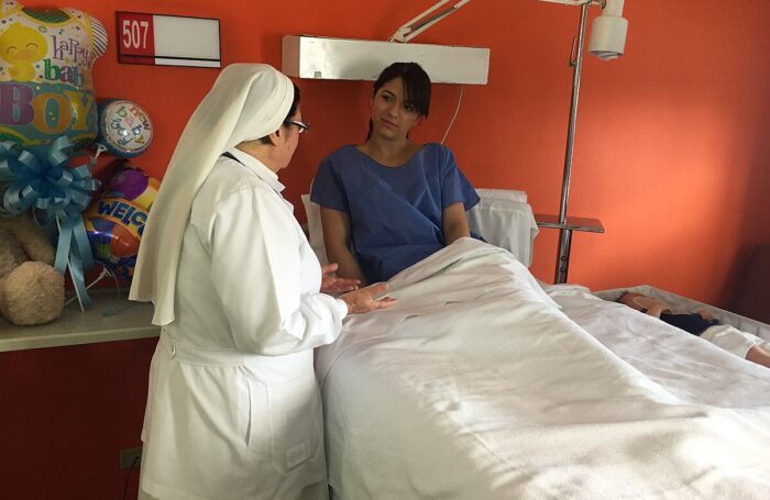 Eine Nonne steht neben einem Krankenhausbett, darin eine Person in einem blauen Hemd