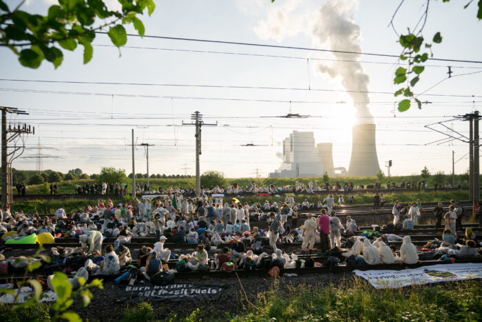 Bild einer Blockade einer Eisenbahnlinie. Mehrere Personen sitzen entspannt auf mehreren Gleisbetten. Im Hitergrund ein Kraftwerk, aus dem Rauch kommt.