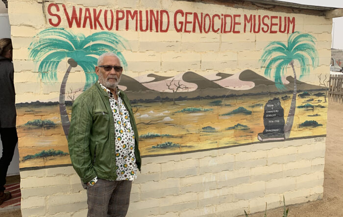 Israel Kaunatjike steht vor einer benalten Wand, die Palmen, eine trockene Landschaft und im Hintergrund Hügel zeigt. Oben steht die Aufschrift "Swakopmund Genocie Museum"