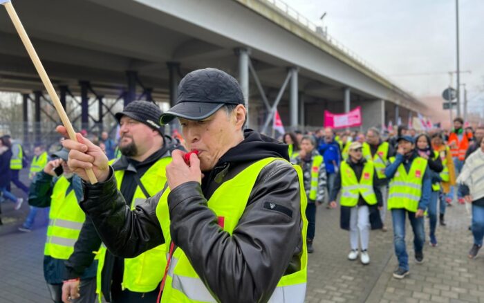 Ein Mann in gelber Warnweste bläst in eine Trillerpfeife, dahinter weitere Demonstrant*innen mit Warnweste unter einer Autobahnbrücke