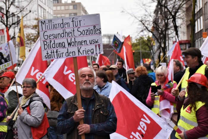 Demonstration mit ver.di Fahnen, im Vordergrund hält ein älterer Mann mit beiden Händen ein Schild hoch. Aufschrift: Milliarden für Gesundheit, Soziales, Bildung, Wohnen etc statt für den Krieg!!