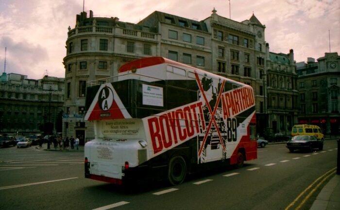 Ein Doppeldeckerbus mit einem bunten Aufdruck: Boycott Apartheid. Dahinter Häuser im viktorianischen Stil.