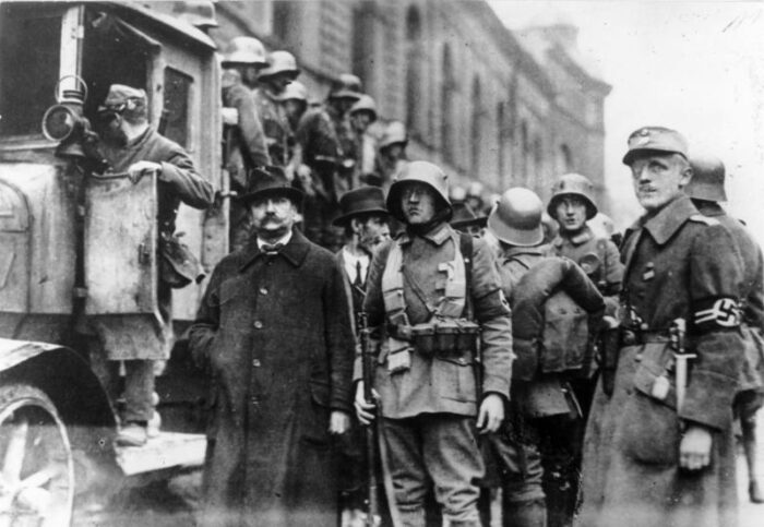 Eine Gruppe bewaffneter Männer, einer mit Hakenkreuzbinde am Arm, in Uniform, daneben ein Mann im Anzug, dahinter ein LKW mit behelmten und bewaffneten Männern.
