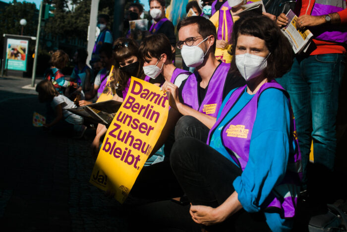 Mehrere Menschen mit Masken und lila Westen sitzen auf der Straße und halten ein PLakat hoch, auf dem steht: "Damit Berlin unser Zuhause bleibt"