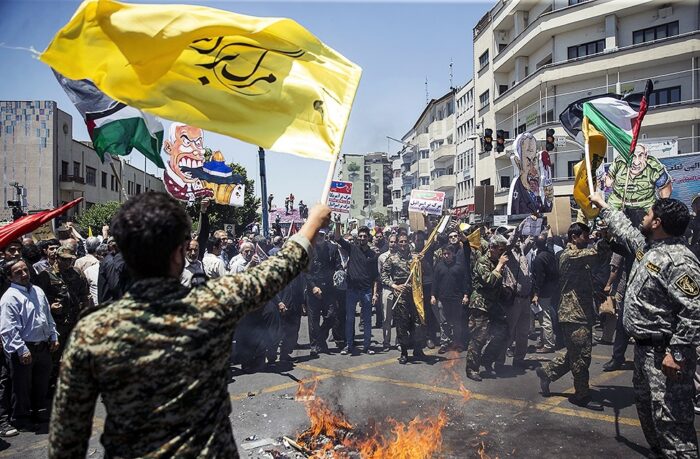 Eine teils uniformierte Menschenmenge steht um ein Feuer auf einer Straße, es werden Fahnen geschwenkt, unter anderem die Palästina-Fahne.