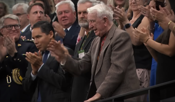 Ein alter Mann im braunen Anzug steht in einer Menge und hebt die Hand zum Winken, die Umstehenden applaudieren