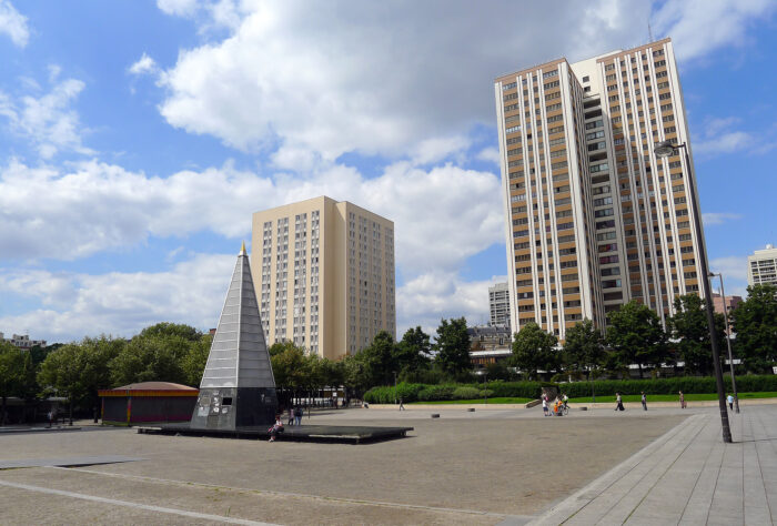 Bild eines Platzes umgeben von hohen Wohnungsbauten und Bäumen. In der Mitte ein Bau, das aussieht wie eine Pyramide aus glas.