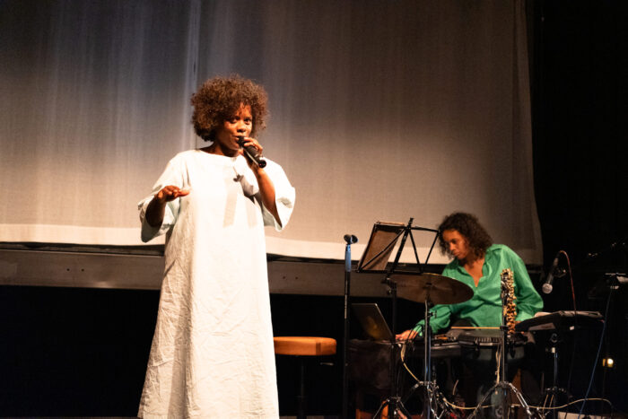 Foto einer Schwarzen Frau in einem weißen Nachthemd, die in ein Mikrofon singt. Die Person steht auf einer Bühne. Im Hintergrund ist eine weitere Schwarze Person.